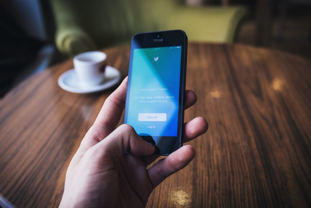 A person's hand holding a smartphone with Twitter on the screen in front of a table with a cup of coffee on it.