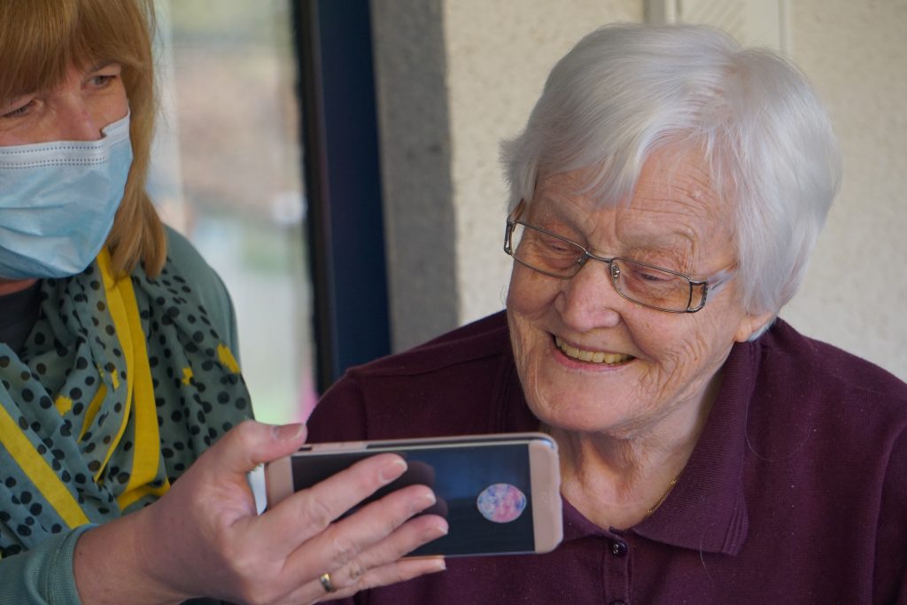 An older woman looking at a smartphone being held by a younger woman wearing a mask